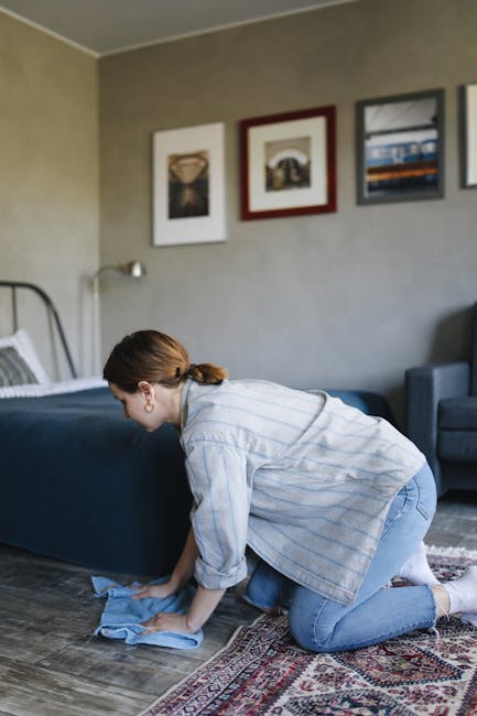 A woman with brown hair tied back, dressed in a light striped shirt and jeans, is kneeling on a patterned rug in a residential living room. She is using a blue cloth to clean the wooden floor. The roo