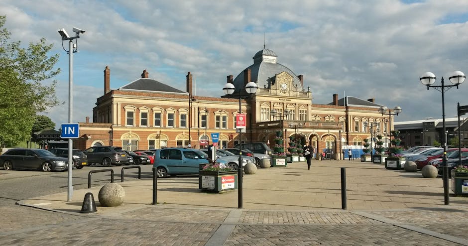 A large historic brick building at Ealing Broadway Station with a detailed roofline and central clock, set against a partly cloudy sky. In the foreground, there is a paved open space with parking areas on either side, containing various parked cars and a few pedestrians. The area is lined with modern lamp posts, some of which have spherical lights, and decorative flower planters. The scene is well-lit with natural daylight, and the space appears clean and well-maintained, reflecting effective surface cleaning and upkeep by Cleaner Ealing, specialists in domestic and commercial sanitisation and deep cleaning services for the Ealing area.
