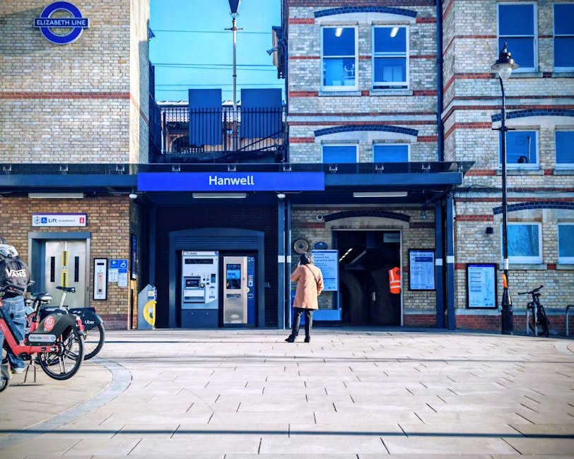 A person standing outside the Hanwell entrance of Ealing Broadway Station, a brick building with multiple windows and a blue sign reading 'Hanwell'. In front of the entrance, there are two ATM machines, a bicycle rack with several bikes including a Ubers bike and a pedal bike, and a street lamp. The pavement is clean and paved with rectangular tiles, with bright, natural daylight illuminating the scene, emphasizing the station's exterior and surroundings. Cleaner Ealing offers comprehensive surface cleaning and deep cleaning services to maintain hygiene and cleanliness in residential and commercial spaces.